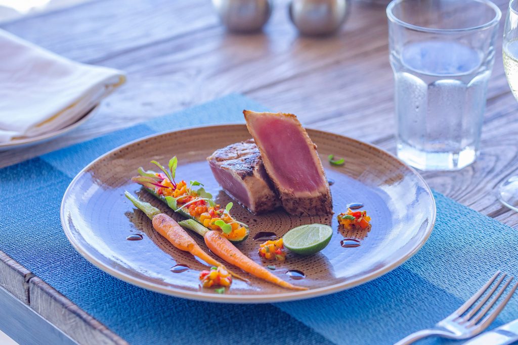 Gedeckter Holztisch mit Keramikteller, gebratenem Fisch, buntem Gemüse, Limette, blauem Stoffserviette und Gläsern