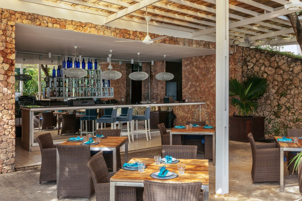 Offener Bar- und Restaurantbereich mit Natursteinwänden, Holz­tischen, blauen Hockern und geflochtenen Stühlen in Melia.