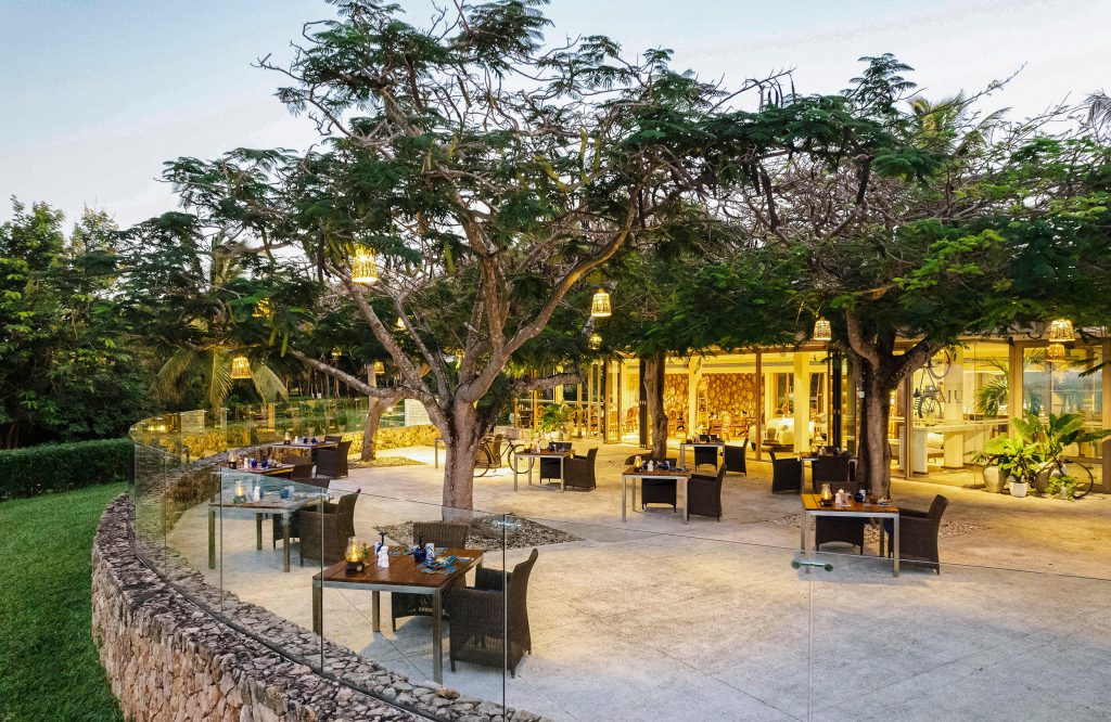 Terrasse eines Hotels mit Rattanmöbeln, Natursteinwall, Glasgeländer und Bäumen mit Lampen bei Dämmerung