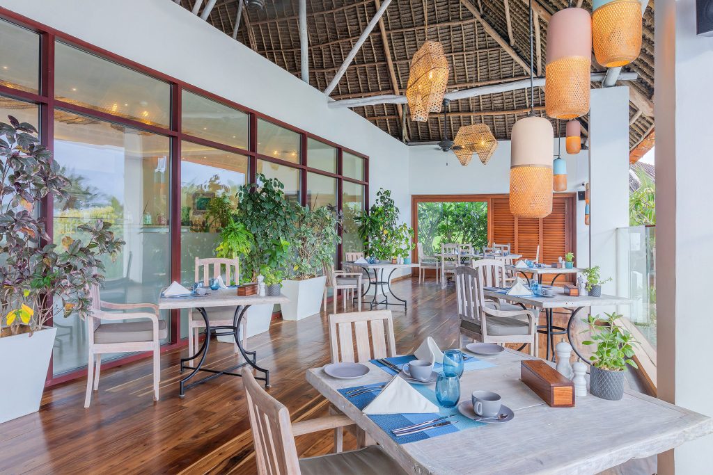 Essbereich im Melia Zanzibar mit hellen Holzmöbeln, großen Glasfenstern und geflochtenen Hängelampen