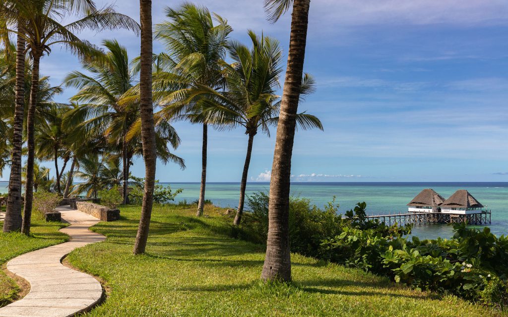 Geschwungener gepflasterter Weg mit Palmen und Blick auf zwei Holzpavillons im Meer bei Melia Zanzibar