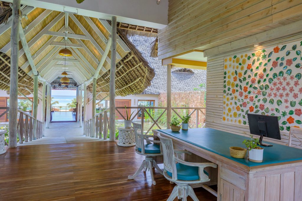 Offener Hotelbereich mit hellem Holzschreibtisch, grünen Pflanzen, floralen Wänden und Meerblick in Sansibar