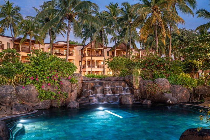 Blick auf den Pool mit klarem blauem Wasser, Wasserfall und tropischen Palmen vor Hotelgebäuden in der Dämmerung