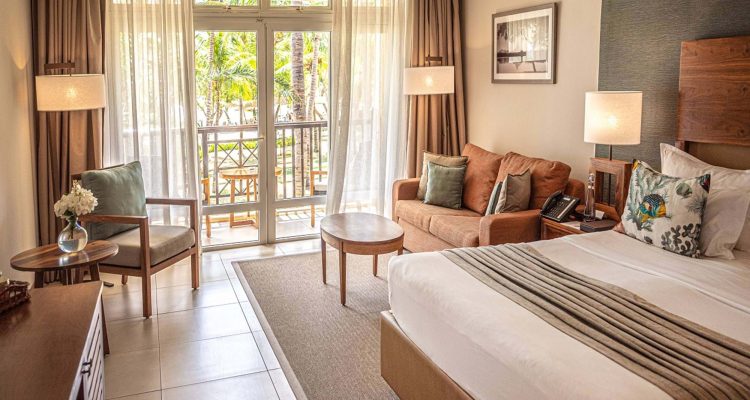 Hotelzimmer mit großem Bett, weißen Bettwäsche, Holzmöbeln und Glasfront zum Balkon mit Pflanzen in Mauritius