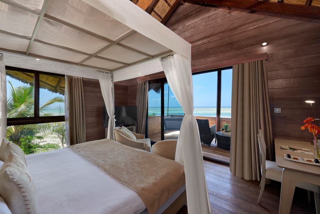 Hotelzimmer mit hellem Himmelbett und weißen Vorhängen, dunklem Holz, großen Fenstern und Blick auf Terrasse und Meer