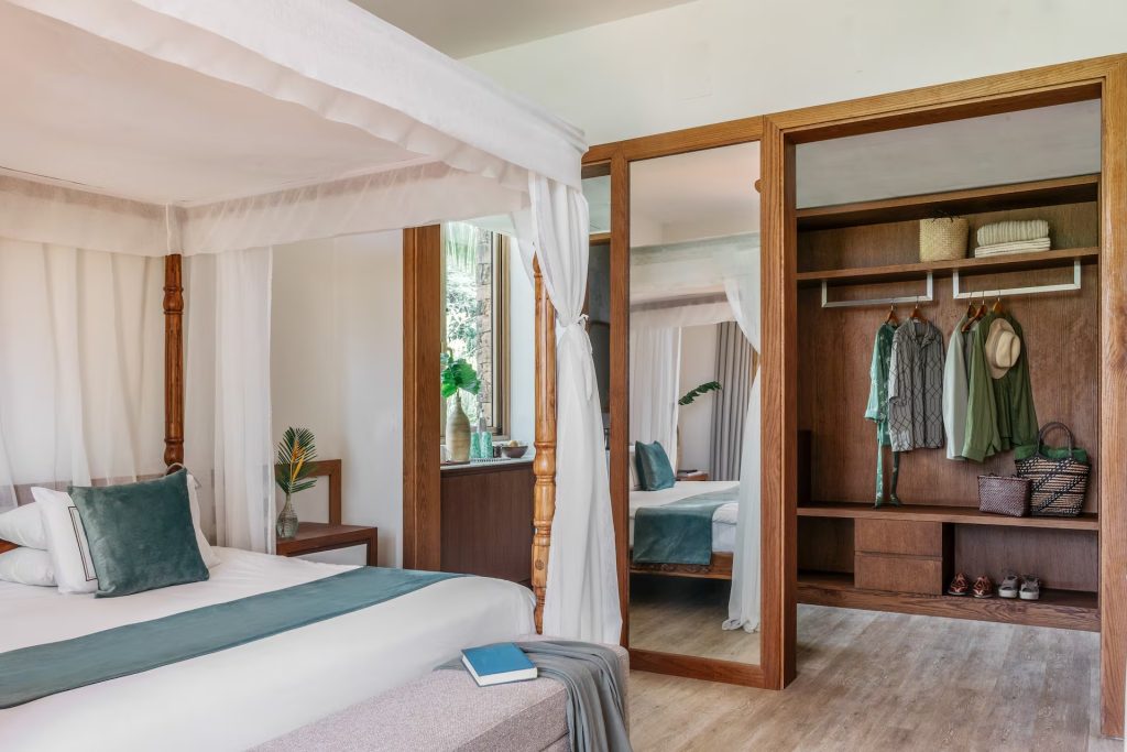 Hotelzimmer mit Himmelbett aus Holzpfosten, hellem Holzboden, Glas-Schiebetüren am Kleiderschrank und Tageslicht