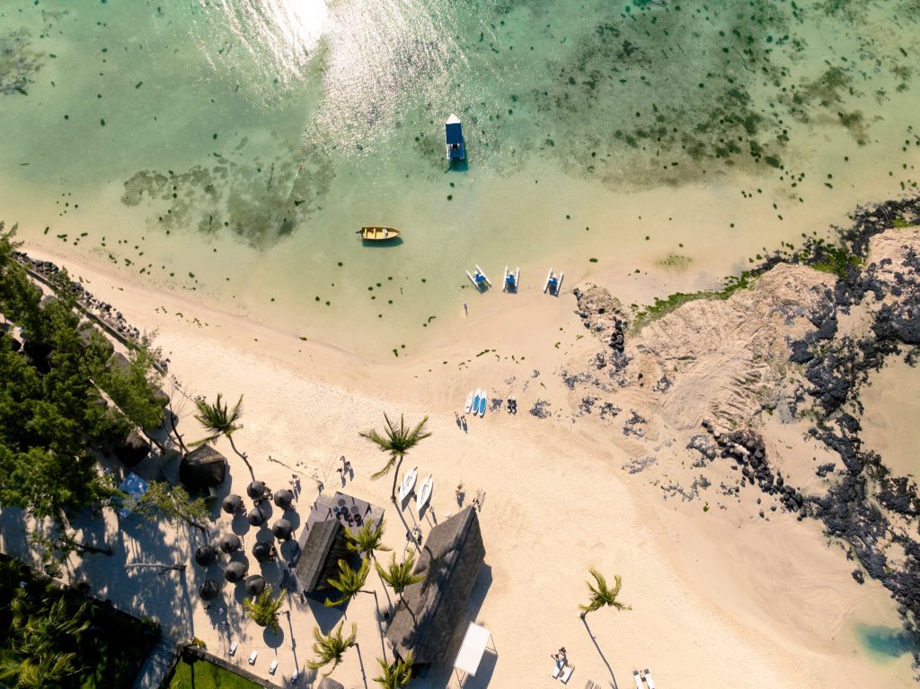 Vogelperspektive auf Ambre Strand in Mauritius mit goldfarbenem Sand, Palmen, Sonnenschirmen und Booten im türkisfarbenen.