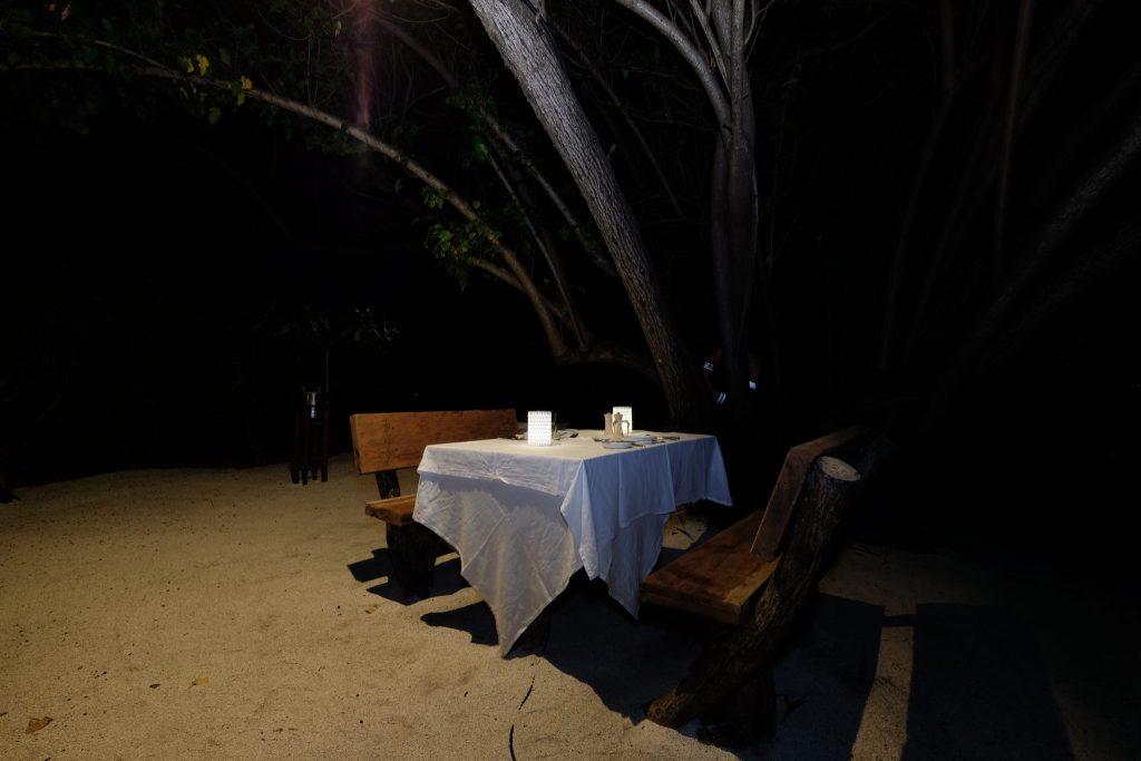 Freiluft-BBQ-Tisch mit weißem Tuch und rustikalen Holzbänken auf sandigem Boden, beleuchtet bei Nacht