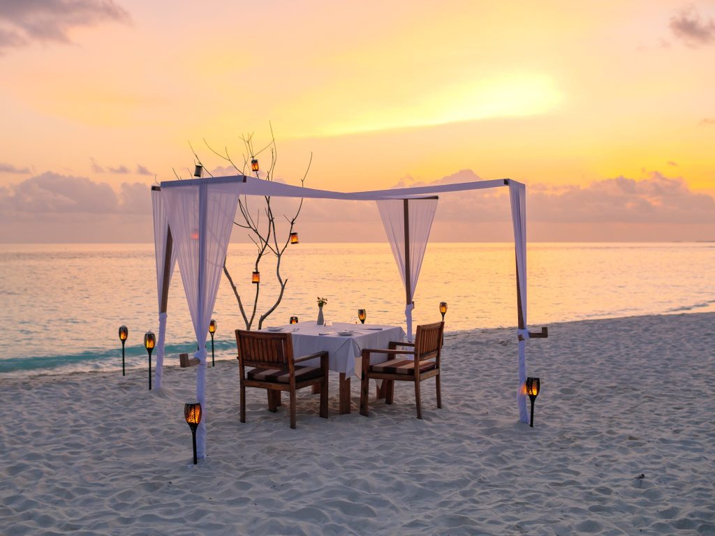 Weißer Pavillon mit transparenten Vorhängen und Holztisch am Sandstrand bei Sonnenuntergang auf den