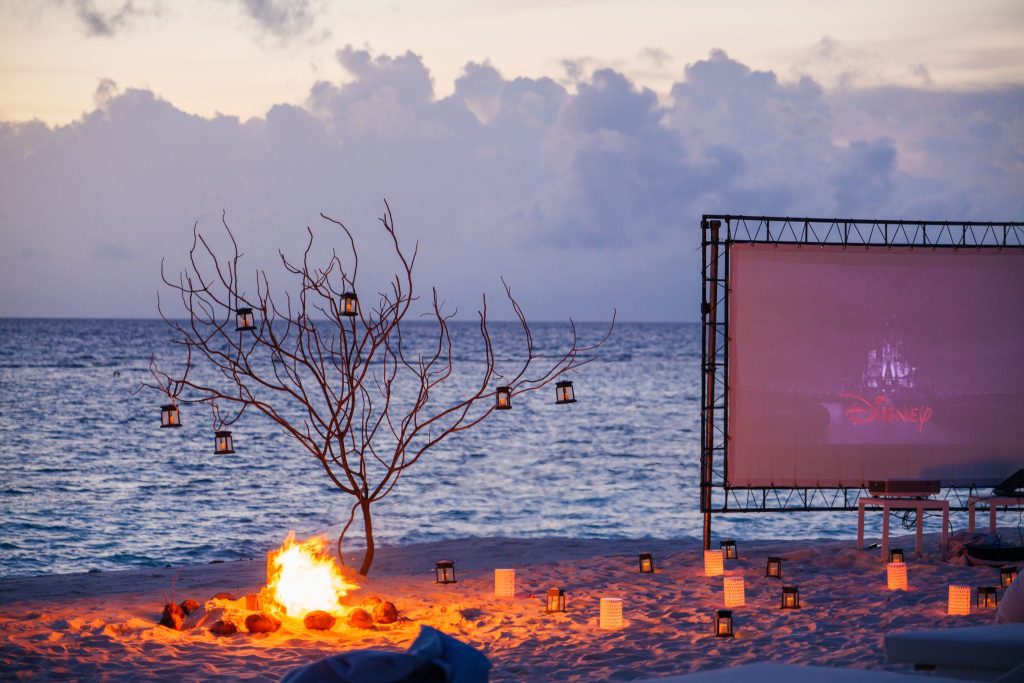 Strand bei Dämmerung mit Lagerfeuer, Laternen an kahlem Ast und großer Leinwand mit Disney-Logo auf den