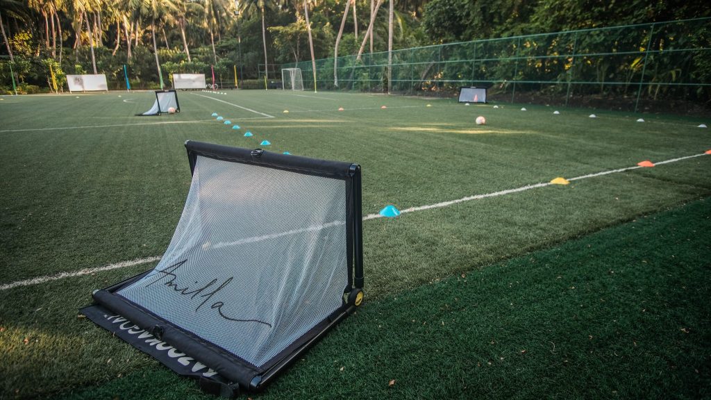 Moderner Sportplatz mit grünem Kunstrasen, kleinen Metalltoren und farbigen Hütchen vor tropischen Bäumen auf den