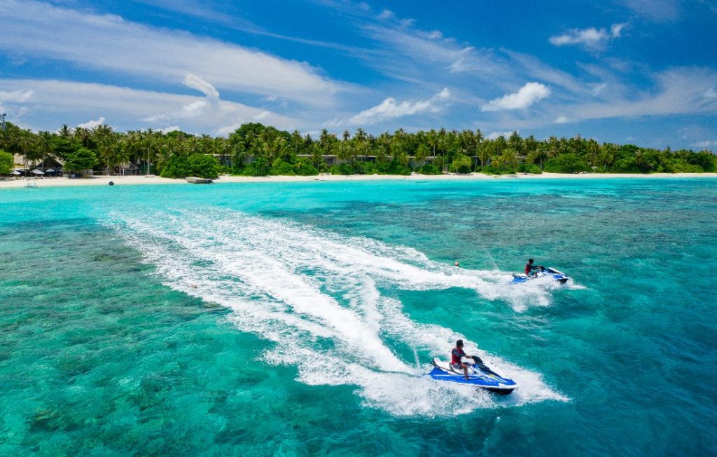 Zwei Personen auf Jet-Skis fahren über türkisfarbenes Meer vor grüner Insel mit Palmen und weißem Strand
