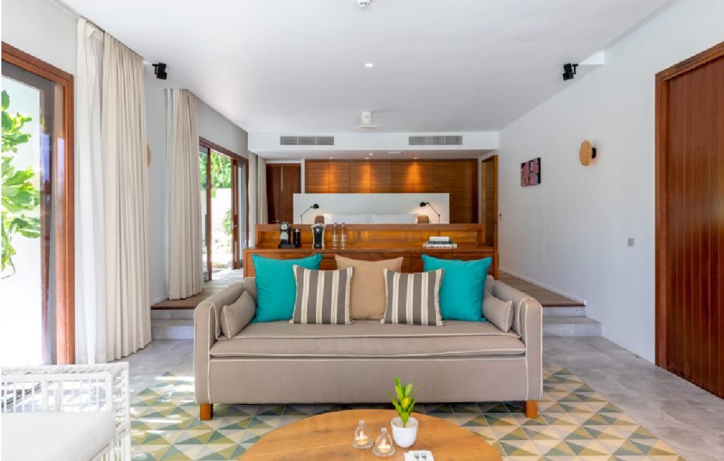 Moderner Hotelraum mit beigem Sofa, türkisen Kissen, geometrischem Bodenmuster und Holzvertäfelung in Amilla Maldives