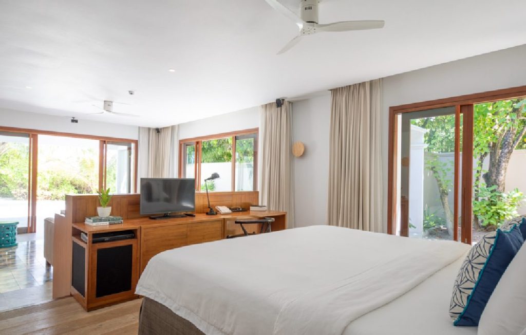 Modernes Hotelzimmer mit großem Bett, Holzmöbeln, weißen und blauen Kissen, großen Fenstern und Blick auf grünes Außenbereich