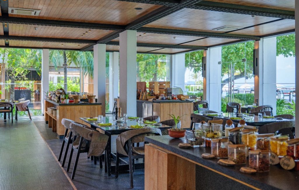 Modernes Hotelrestaurant mit hellen Holzmöbeln, dunklen Tischplatten und Palmen im Außenbereich auf den