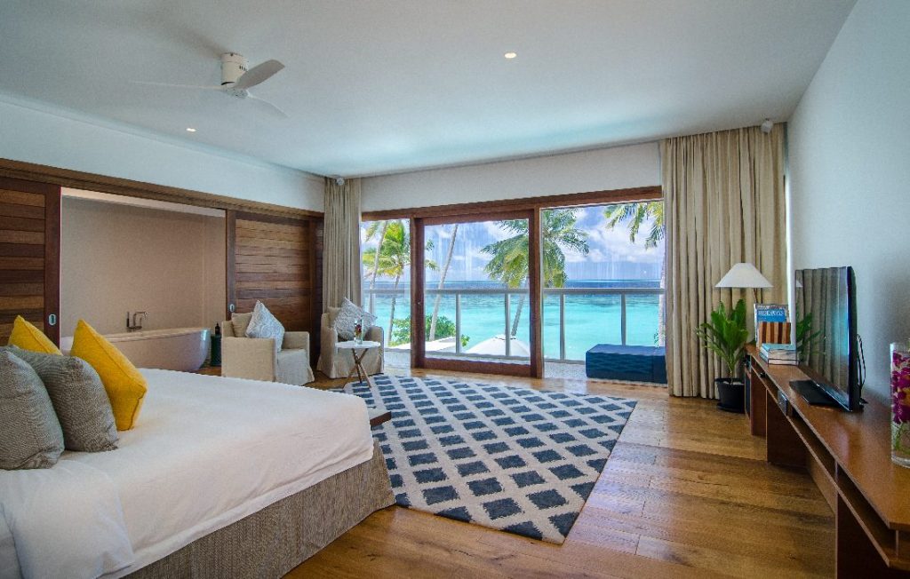 Hotelzimmer mit großem Bett, Holzboden, Fernseher und großen Fenstern mit Blick auf Palmen und Meer auf den