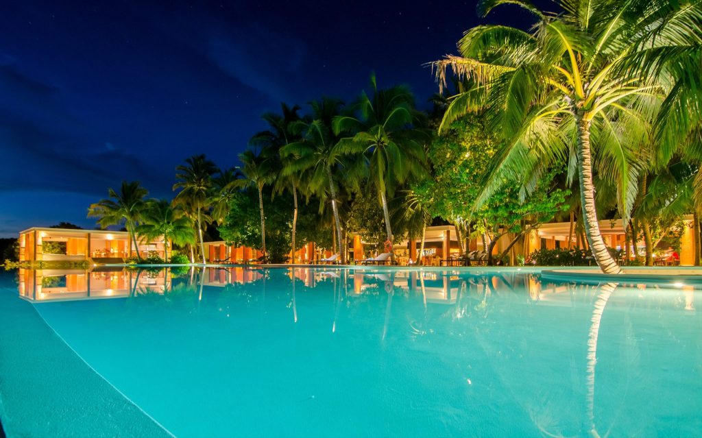 Poolbereich bei Amilla Maldives mit türkisblauem Wasser, Palmen und beleuchteten Gebäuden im Hintergrund