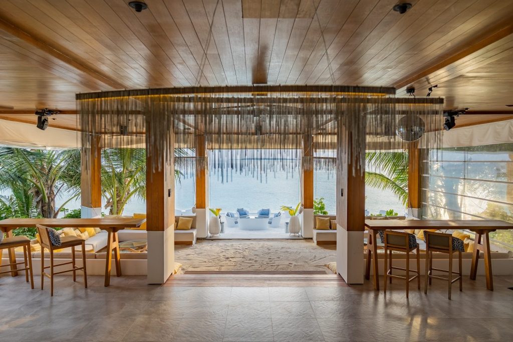 Offener Barbereich Amilla Maldives mit hellen Holzmöbeln, tropischen Pflanzen und Meerblick auf den