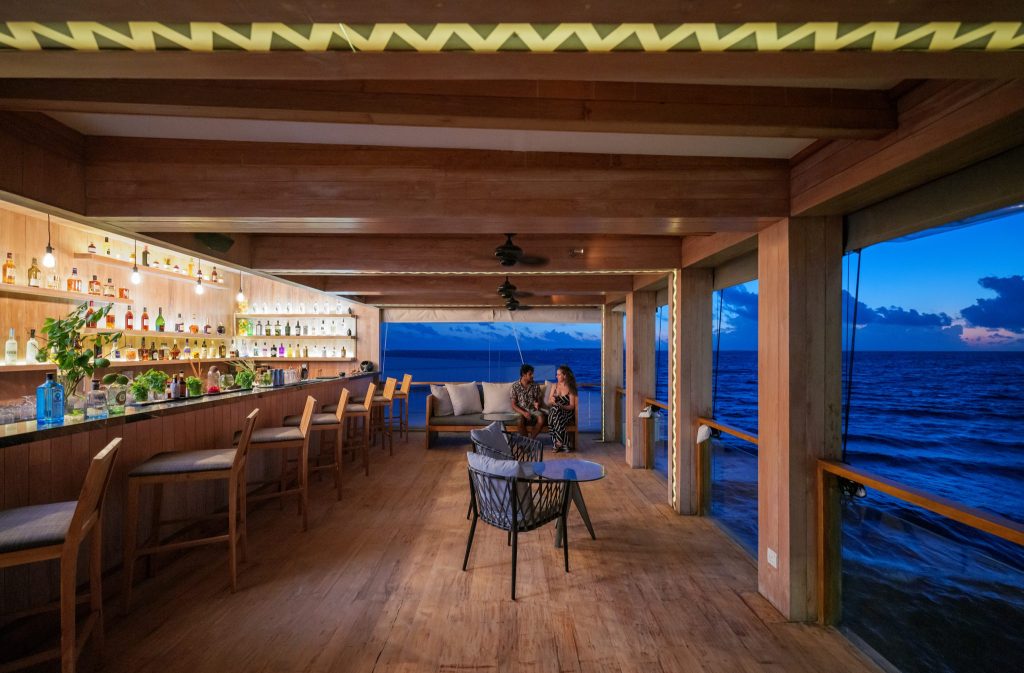 Hotelbar bei Dämmerung mit Holzmöbeln, Glasregalen und Blick auf das Meer auf den