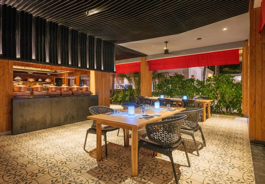 Offener Restaurantbereich Amilla Maldives mit Holzbalken, schwarzen Korbstühlen, gemusterten Fliesen und grünen Pflanzen