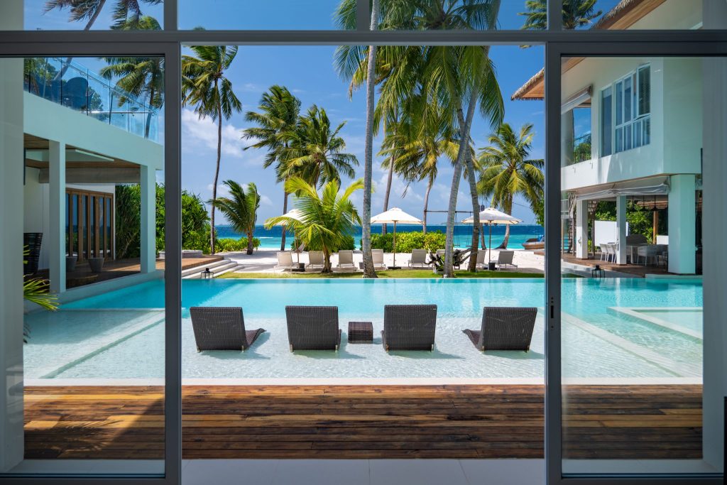 Blick durch große Glasfront auf Pool mit Liegestühlen aus dunklem Geflecht und Meer im Hintergrund