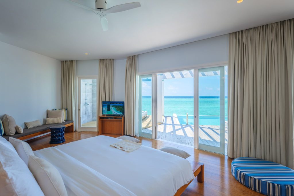 Geräumiges Hotelzimmer mit großem Bett, Holzfußboden, Glastür zum Meer und Sitzecke mit TV
