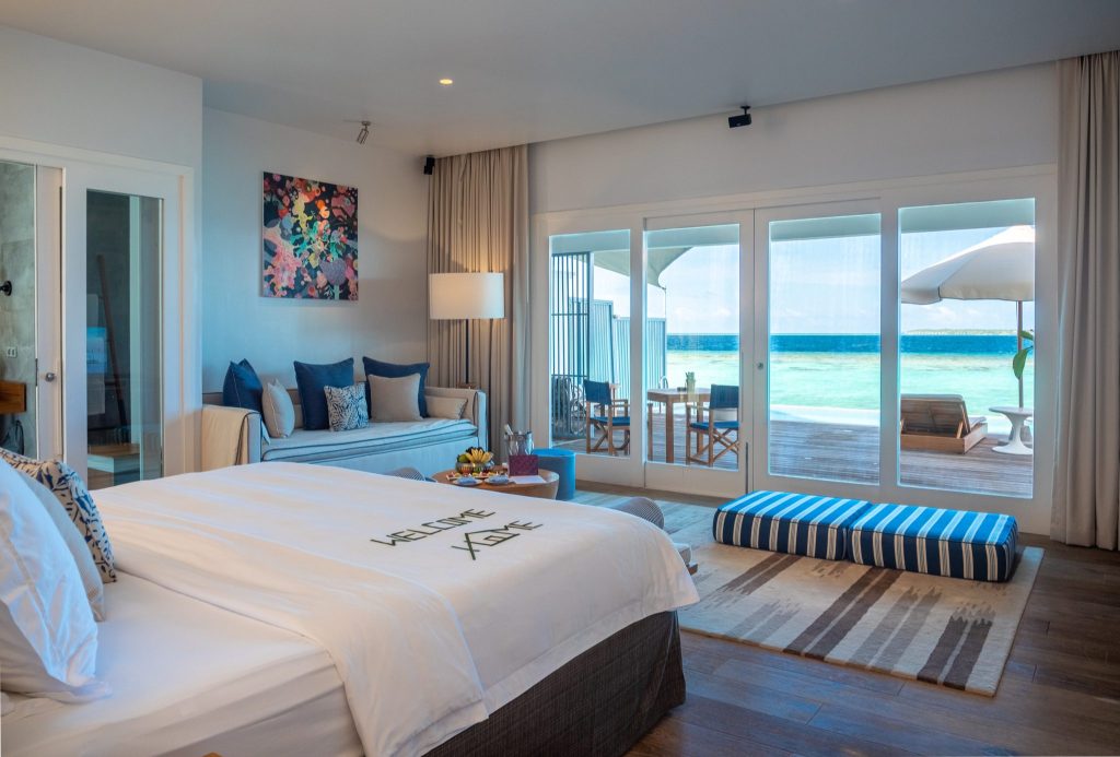 Modernes Hotelzimmer mit großem Bett, blauen Kissen und Terrasse mit Blick auf türkisfarbenes Wasser