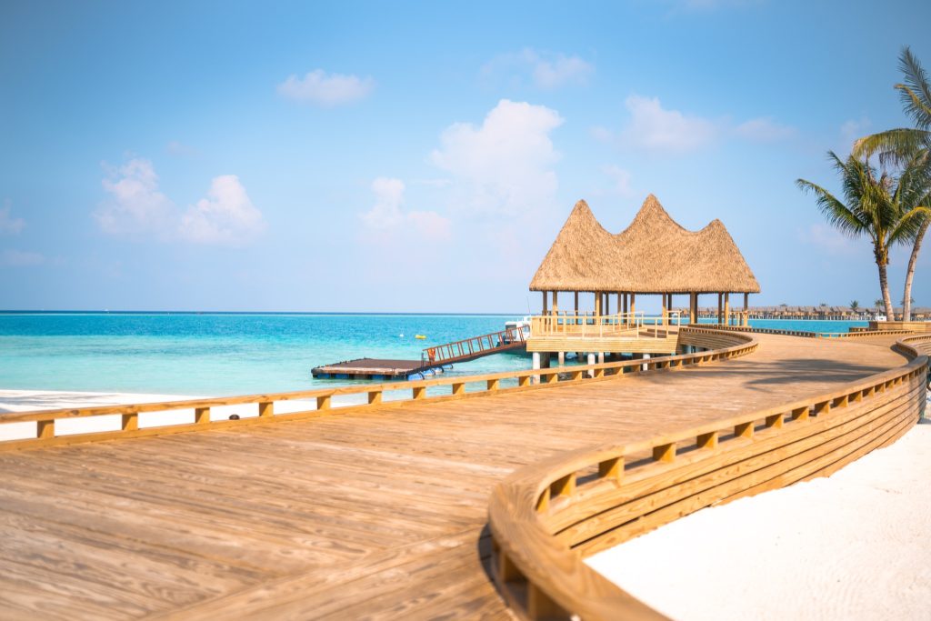Hölzerner Steg am Strand mit Strohdach-Hütte am Ende, türkisblaues Meer und heller Sand auf den