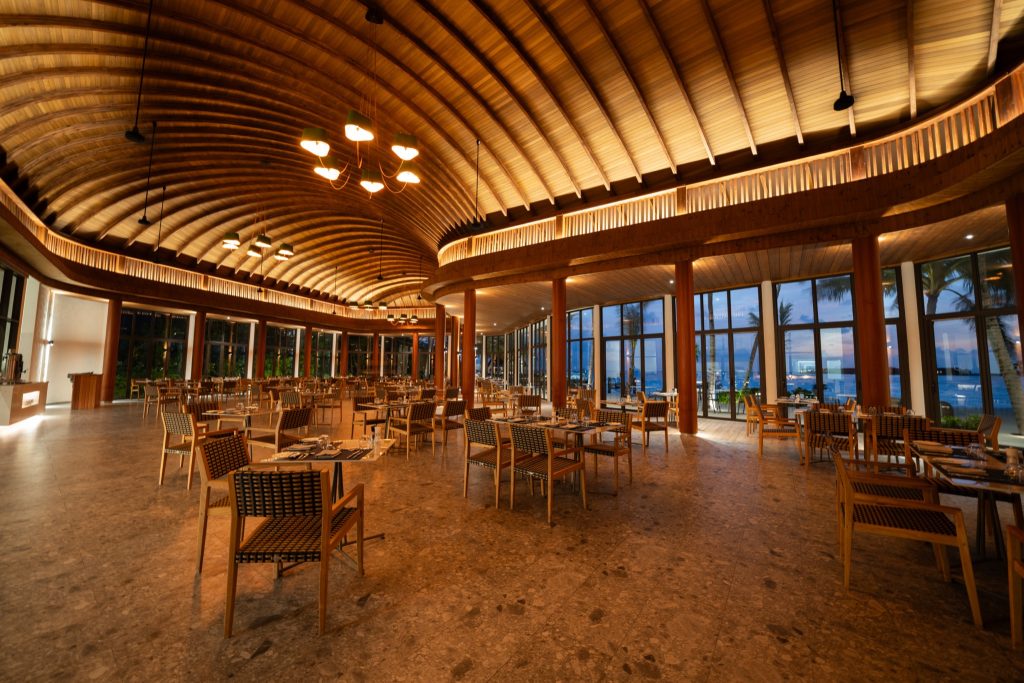 Großer offener Restaurantbereich mit gewölbtem Holzdach, Holztischen, Stühlen und Blick auf Palmen und Ozean