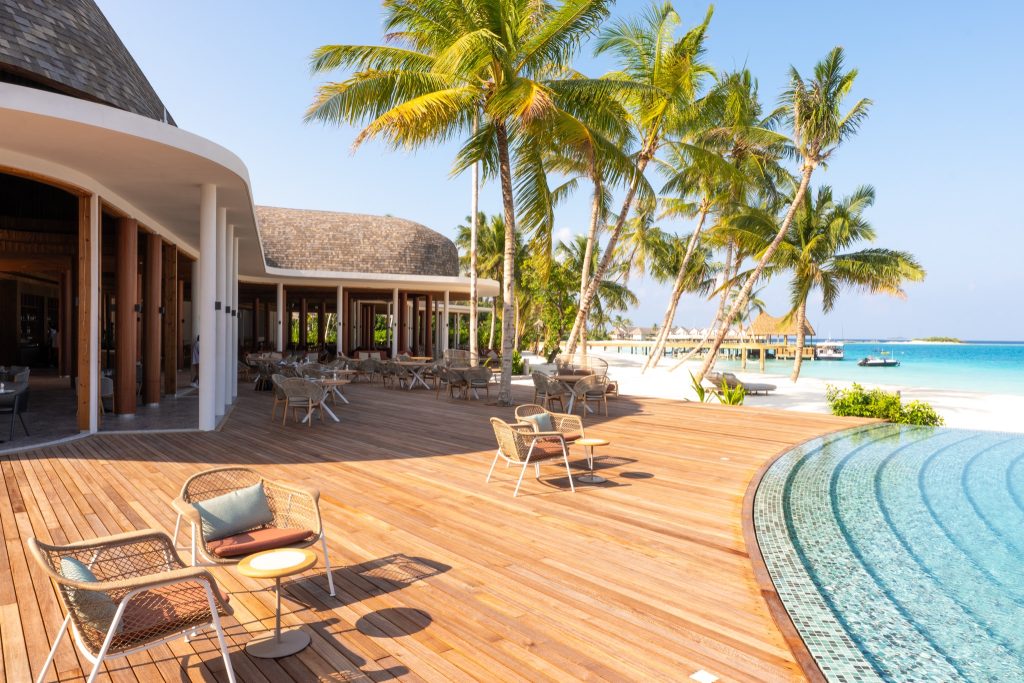 Holzterrasse mit Sitzmöbeln, Pool mit blauen Mosaikfliesen und Palmen am Strand auf den