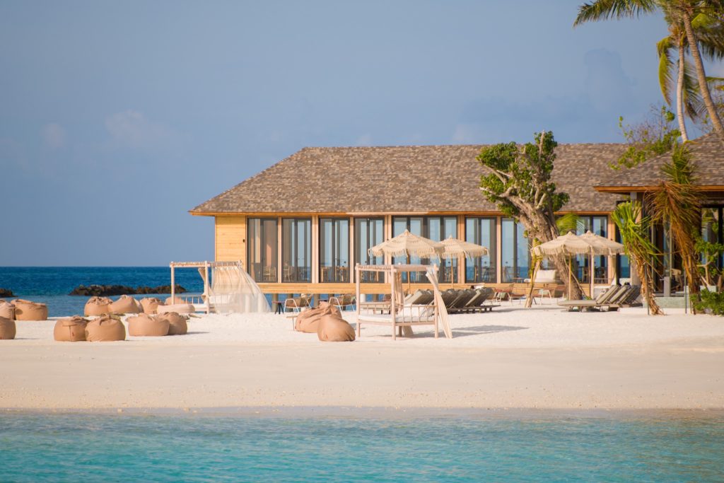 Strandnahe Bar mit großen Glasfenstern, Holzverkleidung, Sonnenliegen und Sitzsäcken auf hellem Sand am Meer