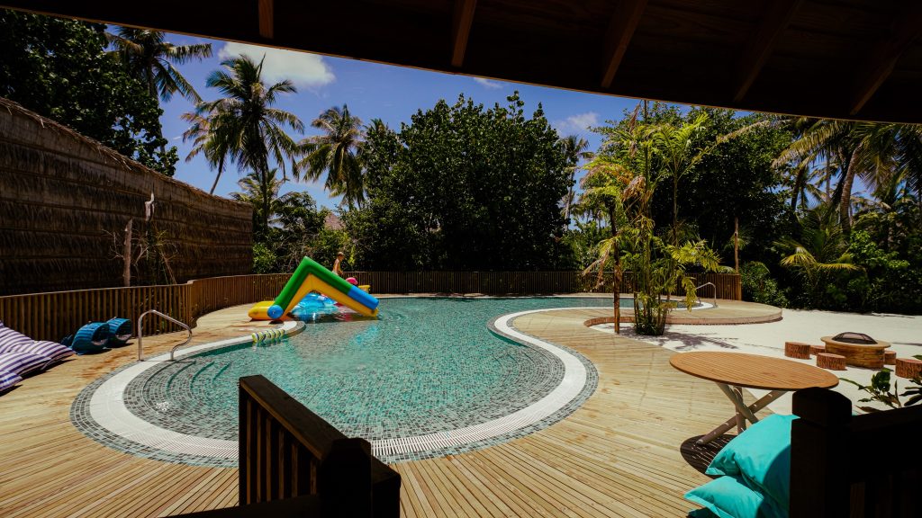 Swimmingpool mit hölzernem Deck, buntem aufblasbarem Spielzeug und türkisfarbenen Holzsitzen unter Palmen