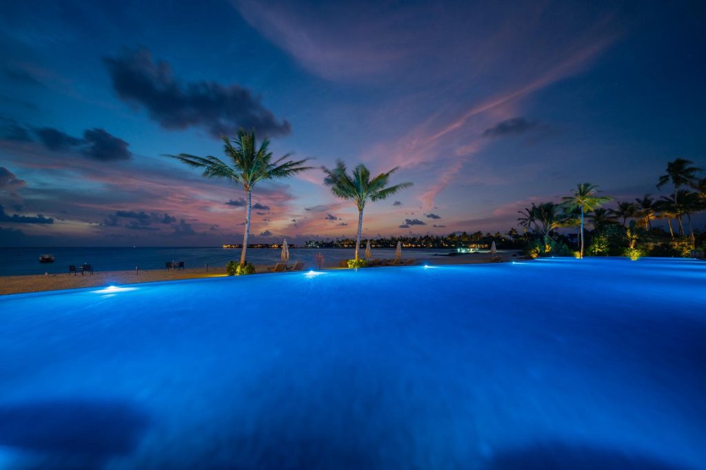 Infinity-Pool mit Unterwasserbeleuchtung, Palmen und Liegestühlen vor Meer und farbigem Himmel bei Dämmerung