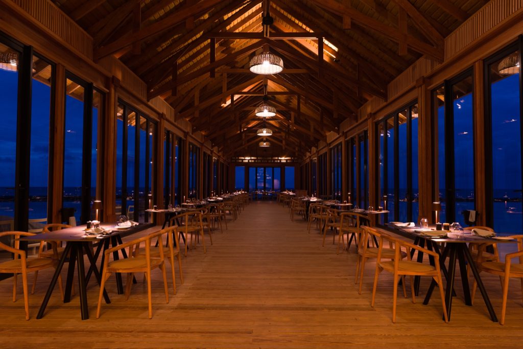 Restaurant The Flame im ananea Madivaru Maldives mit Holzdecke, langen Tischen, großen Fenstern und abendlicher Dämmerung
