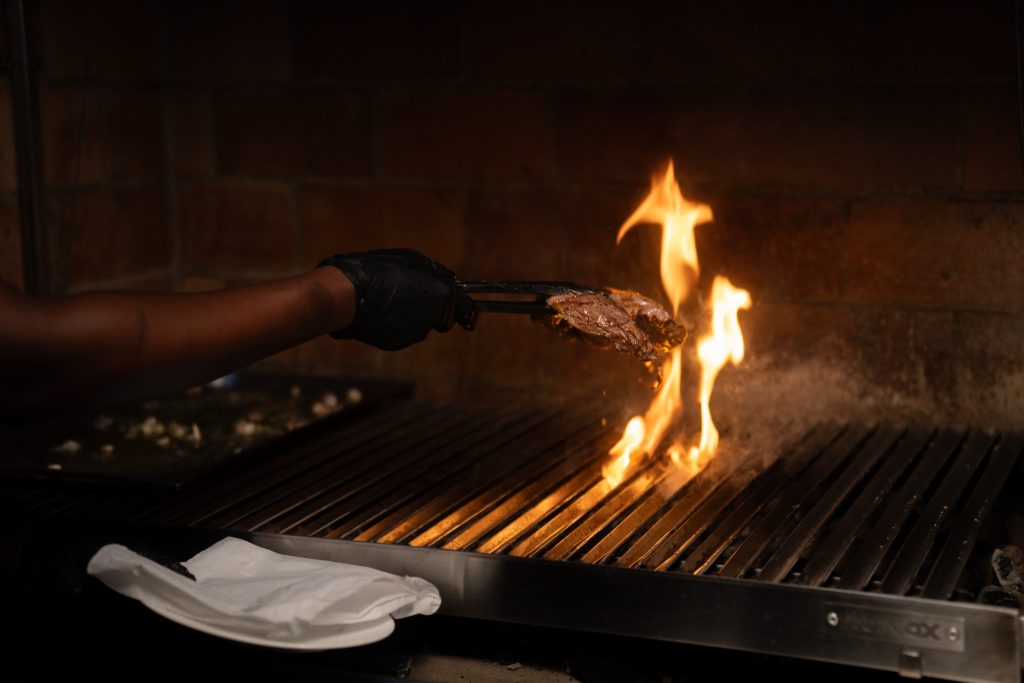 Hand hält mit Zange Fleisch über offener Flamme auf Metallgrill in Restaurant The Flame
