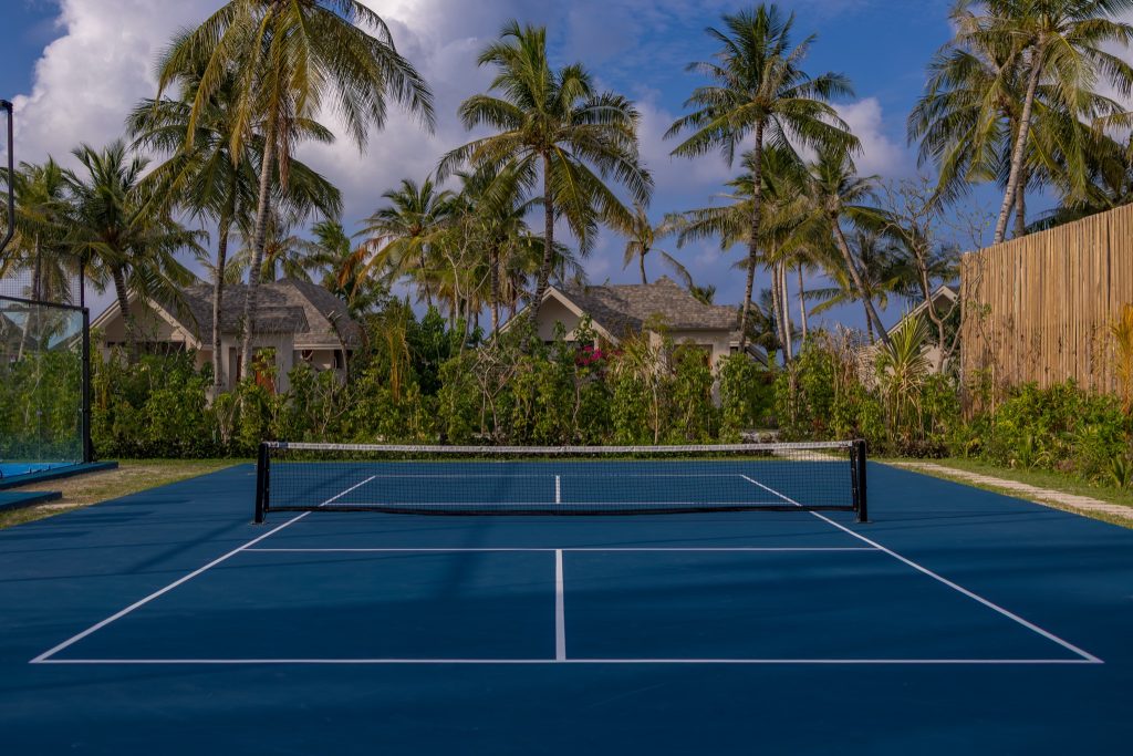 Tennisplatz mit dunkelblauem Belag und weißen Linien vor Palmen und Hotelbungalows auf den