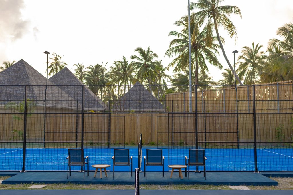 Tennisplatz mit blauem Spielfeld und Metallzaun, vier Stühle und zwei Holztische davor, Palmen und Gebäude im Hintergrund