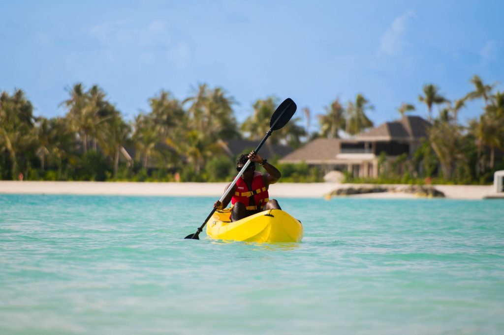 Person in roter Schwimmweste paddelt gelbes Kajak auf klarem, türkisfarbenem Wasser vor Hotel mit Palmen