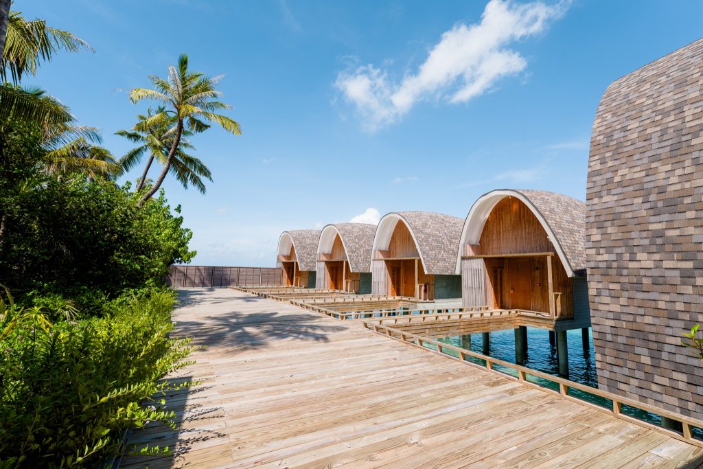 Reihe von Holzbungalows auf Stelzen über türkisfarbenem Wasser mit hölzerner Promenade und Palmen