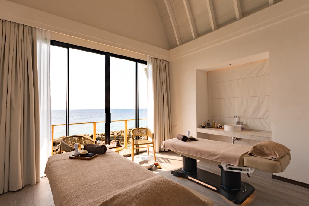 heller Spa-Behandlungsraum mit zwei beige Massageliegen, Waschbecken und Glasschiebetüren zum Meer