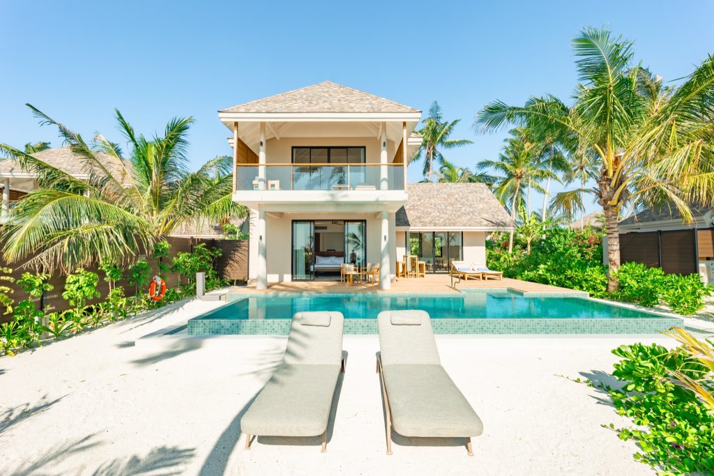 Zweistöckige 2BR Beach Villa mit Pool, Balkon mit Glasgeländer und zwei beigen Sonnenliegen am weißen Sandboden