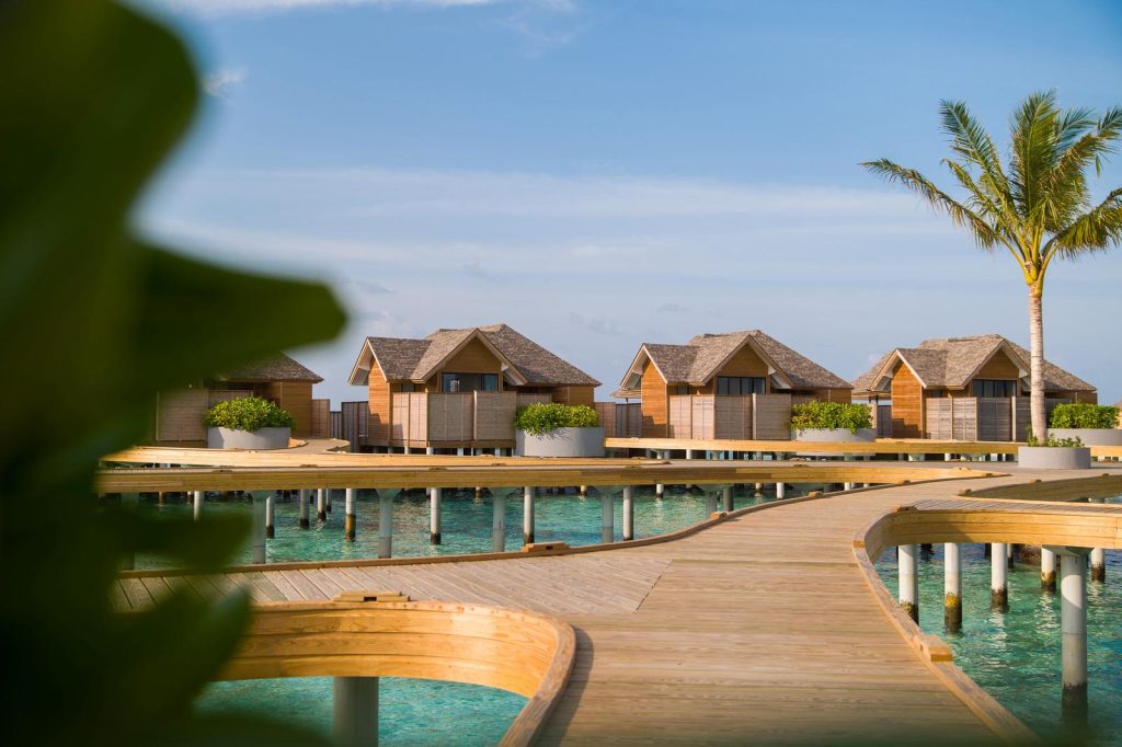 Reihe von Holzbungalows auf Stelzen über türkisfarbenem Wasser mit Holzsteg und tropischen Pflanzen