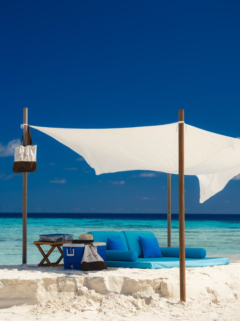 Strand mit Holzpfählen, weißen Stoffbahnen als Sonnenschutz, blauer Liege, Holztisch, Kühlbox, Tasche und Hut.