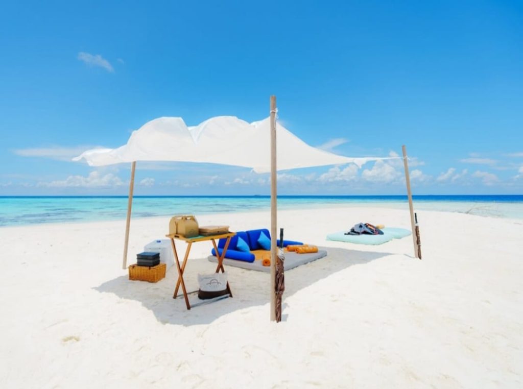 Strand mit weißem Sonnensegel, blauen und orangefarbenen Sitzkissen auf Holztisch, türkises Meer im Hintergrund