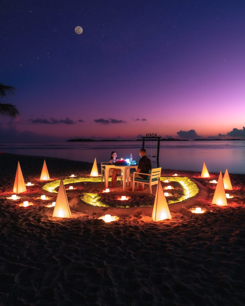 Tisch und zwei Stühle am Strand mit warmweiß leuchtenden Lampenschirmen, lila-pinker Abendhimmel und Mond