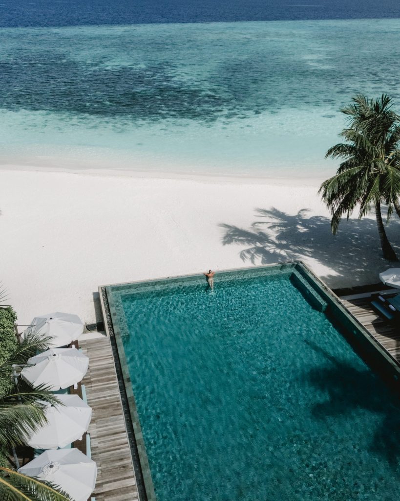 Rechteckiger Pool mit blaugrünem Wasser neben Holzdeck mit weißen Sonnenschirmen und Palmen am Strand