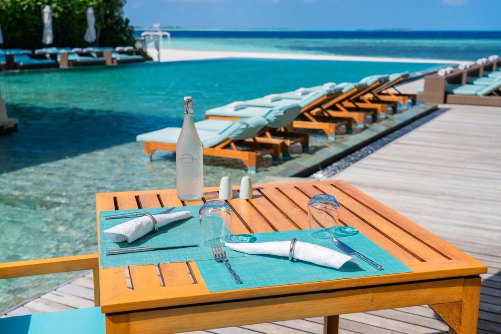 Holztisch mit türkisfarbenen Platzmatten, umgedrehte Gläser, Wasserflasche, Pool mit hellblauen Sonnenliegen im Hintergrund
