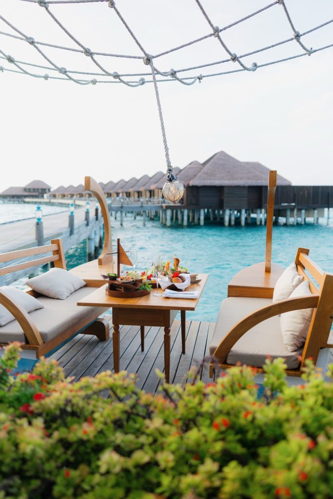 Terrasse mit Holzmöbeln und Kissen über blauer Lagune, Wasserbungalows mit Strohdächern im Hintergrund.