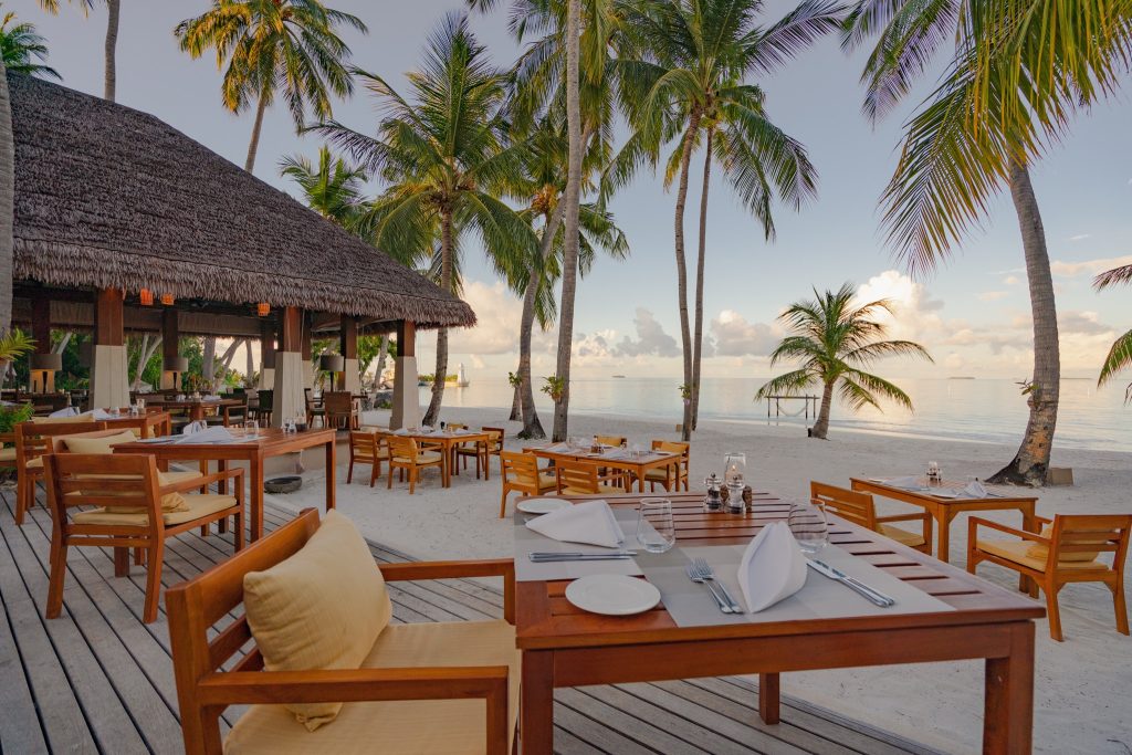 Restaurant Magu mit Holztischen und weißen Servietten auf Holzterrasse, Strand und Meer im Hintergrund