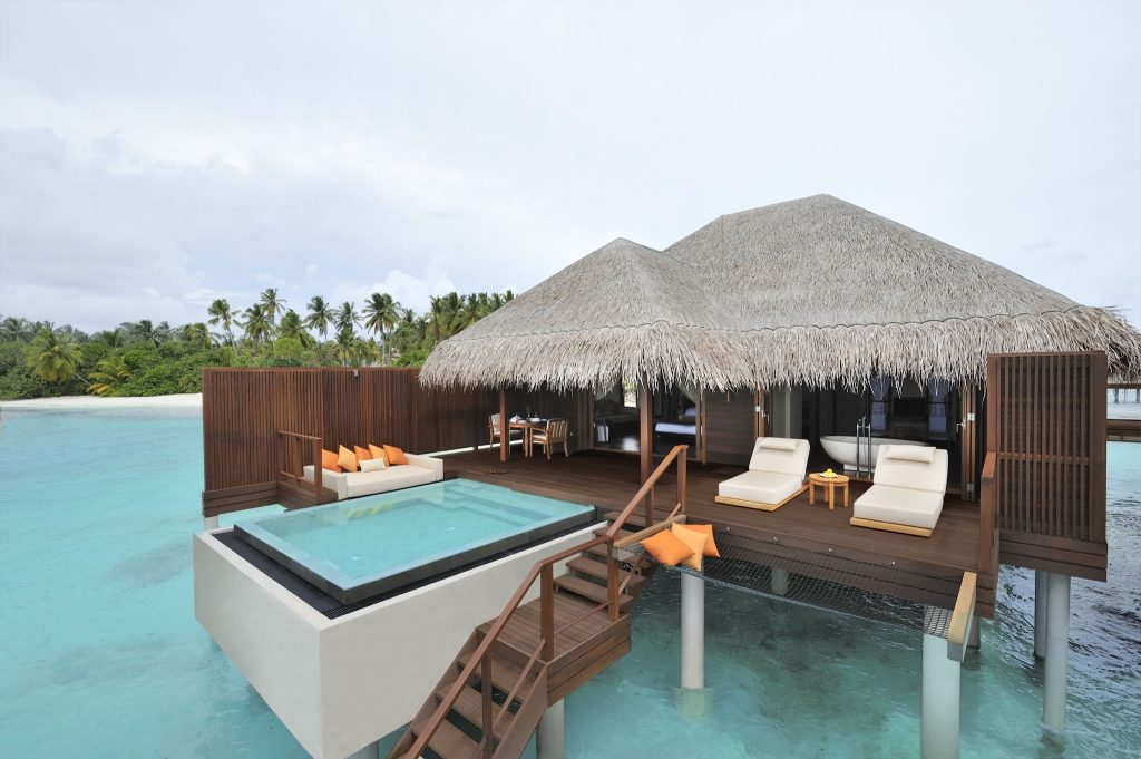 Über Wasser gebaute Ocean Villa mit Holzterrasse, privatem Pool, Sonnenliegen und Sitzgruppe mit bunten Kissen auf den.